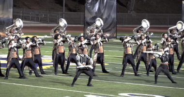 A marching band performs on at Cougar Stadium at College of the Canyons under stadium lights. The brass players, wearing matching ornate jackets and black pants, hold their instruments up in unison while two color guard members in contrasting silver and black outfits strike dynamic poses in front. The scene is highly synchronized and theatrical, with backdrops in the background and a dramatic sense of movement and energy.